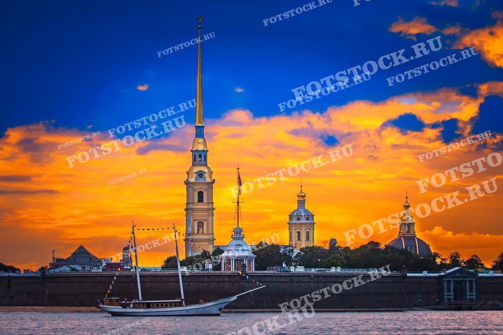 На фотографии изображена Петропавловская крепость в Санкт-Петербурге на фоне яркого закатного неба. Видны собор Петропавловской крепости и корабль на переднем плане. Фото может использоваться для туристических буклетов, открыток и рекламных материалов.